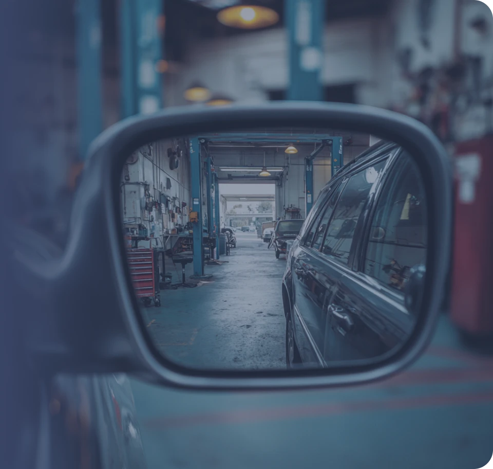A view of a car repair garage seen through a car's side mirror. The mirror reflects the long workshop with car lifts, tools, and equipment, and also shows the dark side of the car the mirror is attached to. The image has a cool, blue-gray filter.