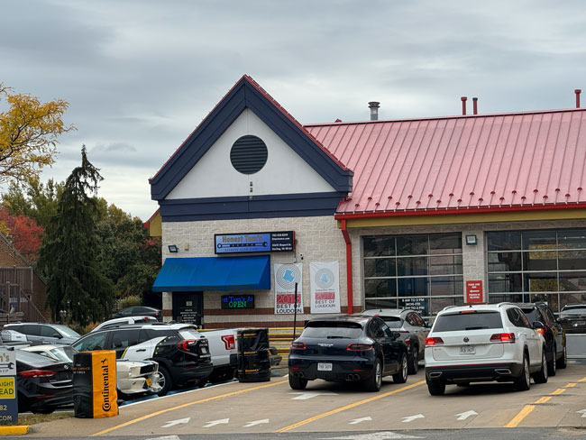 A car repair shop with a red roof and parking lot filled with vehicles. Signs on the building read “Honest Tom’s Auto Care” and “Open.” Trees and cloudy sky are visible in the background.
