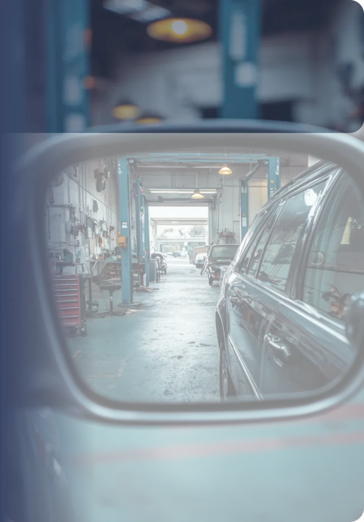 A view of a car repair garage seen through a car's side mirror. The mirror reflects the long workshop with car lifts, tools, and equipment, and also shows the dark side of the car the mirror is attached to. The image has a cool, blue-gray filter.
