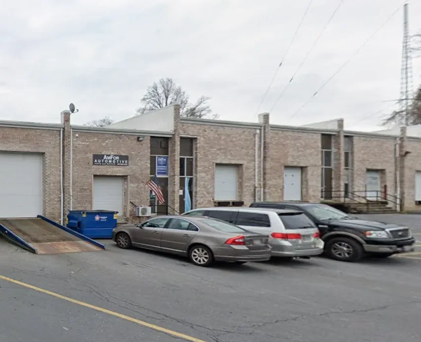 A row of cars is parked in front of a brick auto repair shop with garage doors and a ramp. There’s a sign reading “AutoR Automotive” and an American flag beside the entrance. Trees are visible in the background.
