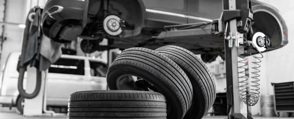 A black and white image of an auto repair shop bay, showing a vehicle raised high on a lift with all its wheels removed, exposing the brake rotors. In the foreground, several new tires are stacked on the floor.