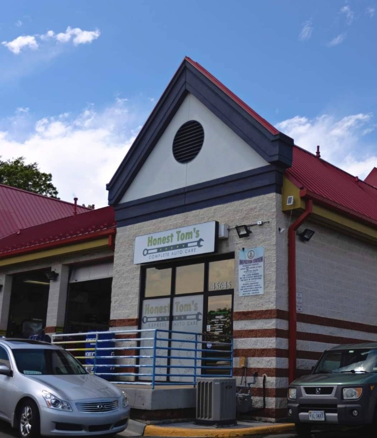 The front of an auto repair shop with light brick siding and a blue awning over the door, displaying a sign for Honest Tom's Complete Auto Care and a large yellow banner that reads Tire Experts.